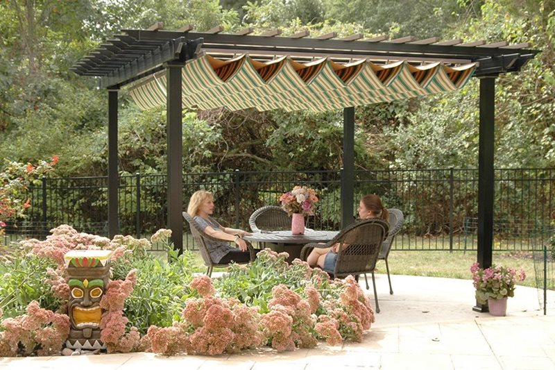 2 people sitting at an outdoor table under a black pergola with a green and tan striped pergola canopy.